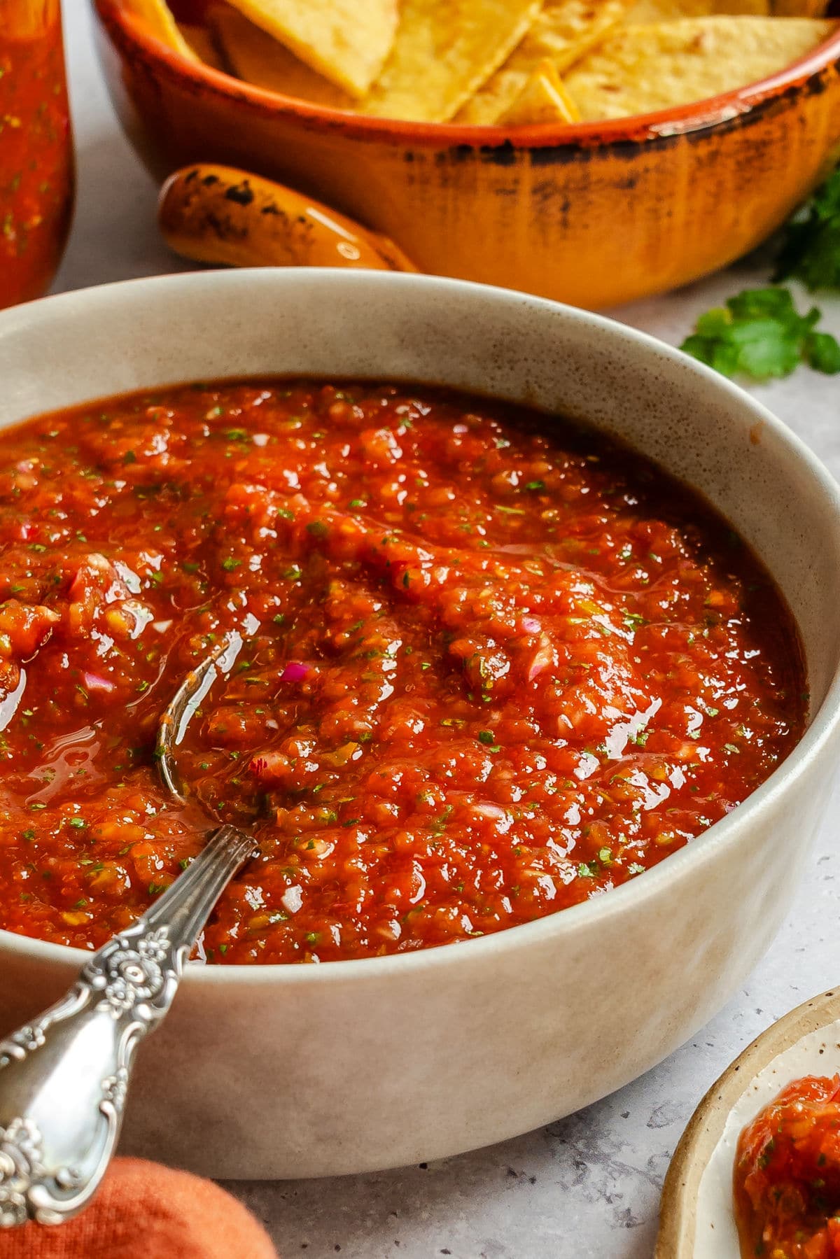 A dish of blender salsa next to a dish of chips and fresh cilantro. 