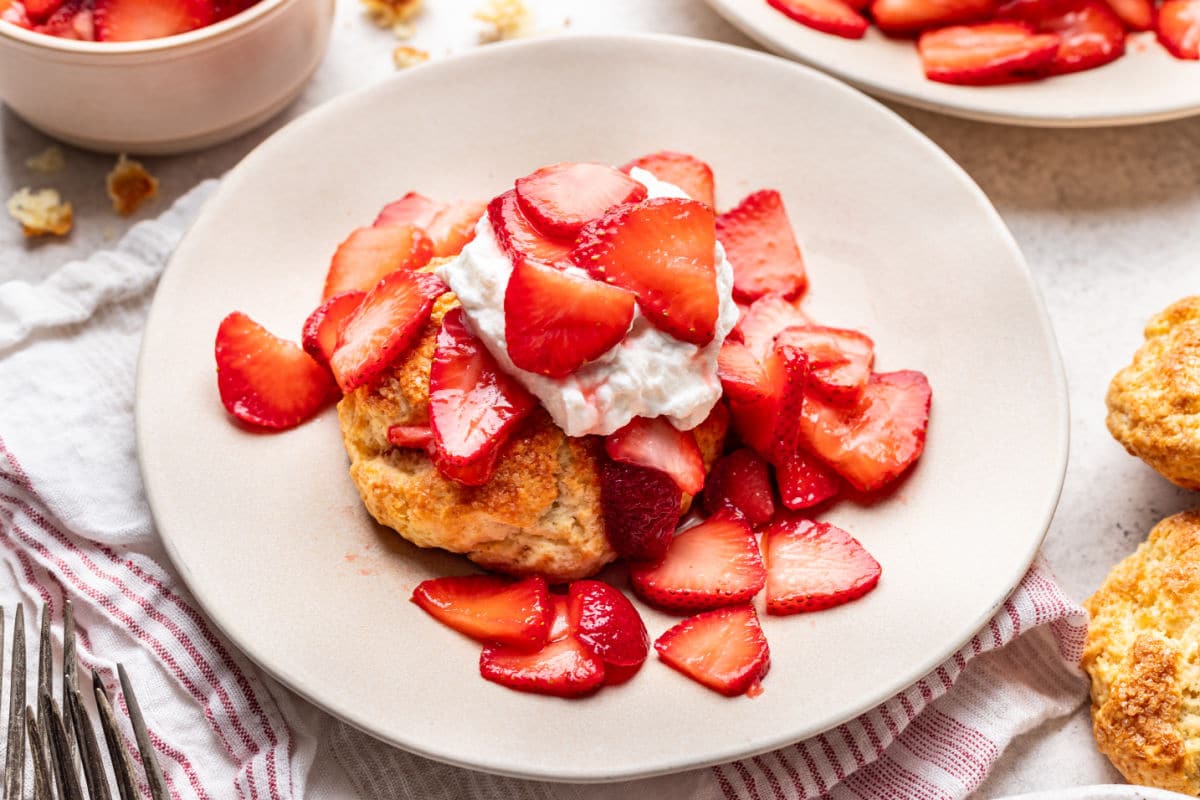 A strawberry shortcake on a plate next to biscuits and a dish of strawberries.