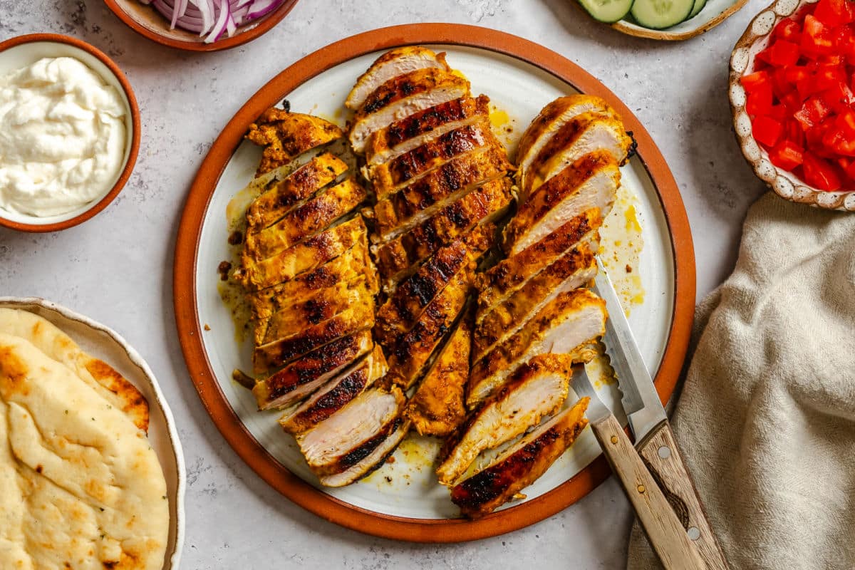 Sliced up chicken shawarma chicken breasts on a plate next to toppings. 