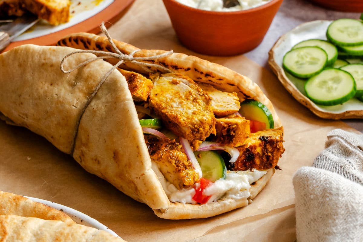 Chicken shawarma in a pita next to sliced cucumber in a dish of toum. 