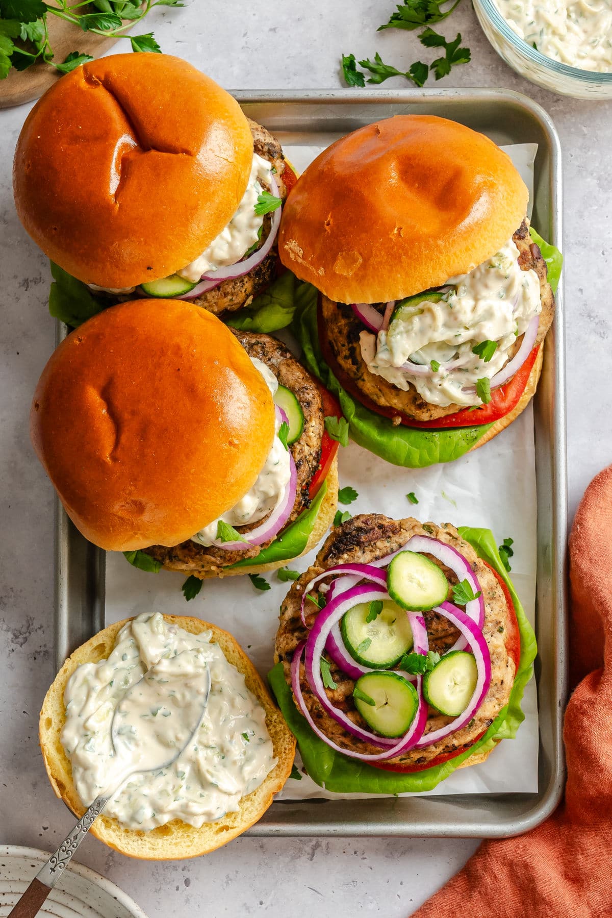 A tray of Greek turkey burgers with one missing its top bun next to a bowl of tzatziki.
