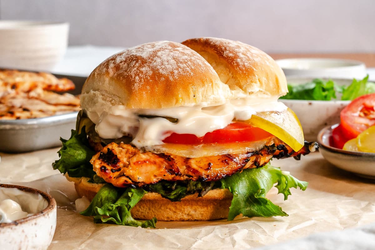 A grilled chicken sandwich next to a dish of tomatoes and pickles. 