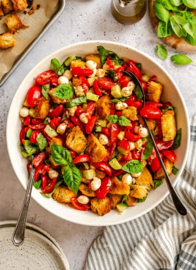 A bowl of panzanella salad next to a kitchen linen and a stack of plates.