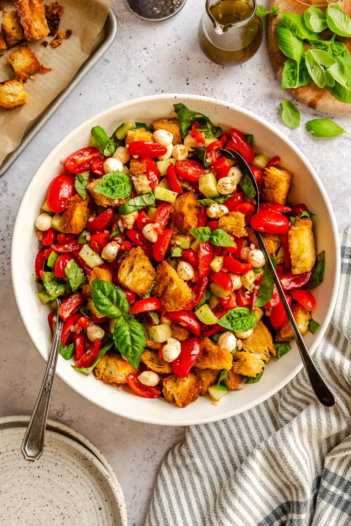 A bowl of panzanella salad next to a kitchen linen and a stack of plates.