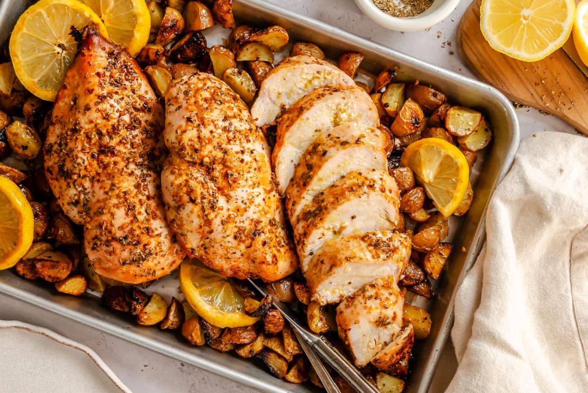 A sliced lemon garlic chicken breast on next to unsliced breasts on roast potatoes. 