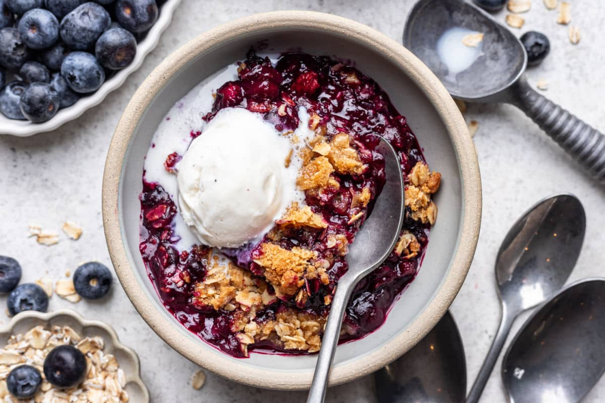 A bowl of blueberry crisp next to an ice cream scoop and a dish of blueberries. 