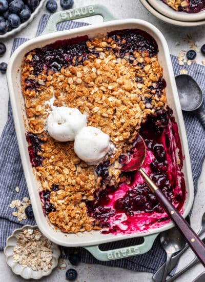 A dish of blueberry crisp topped with two scoops of vanilla ice cream and a spoon in it.