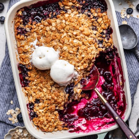 A dish of blueberry crisp topped with two scoops of vanilla ice cream and a spoon in it.