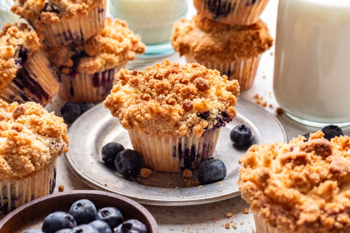 A blueberry streusel muffin on a plate next to a dish of blueberries and a glass of milk.