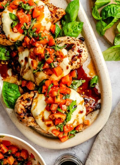 A platter of grilled bruschetta chicken next to bruschetta topping and fresh basil.
