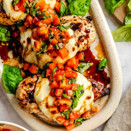 A platter of grilled bruschetta chicken next to bruschetta topping and fresh basil.
