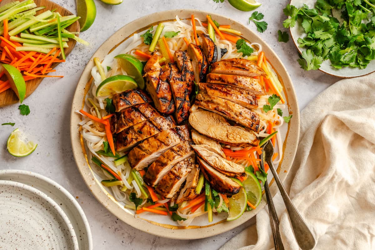 A plate of sliced lemongrass chicken over rice noodles and fresh veggies with two forks under it.