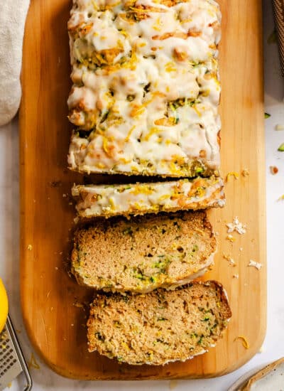 A loaf of lemon zucchini bread partially sliced on a wooden cutting board.