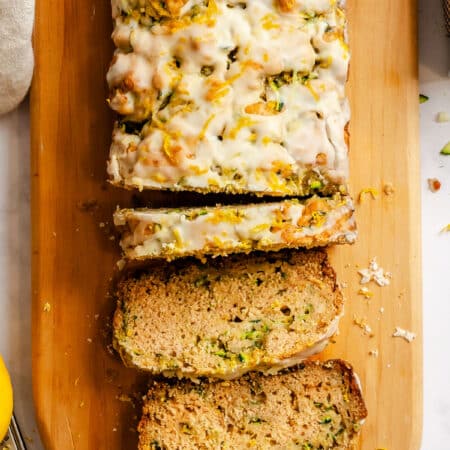 A loaf of lemon zucchini bread partially sliced on a wooden cutting board.