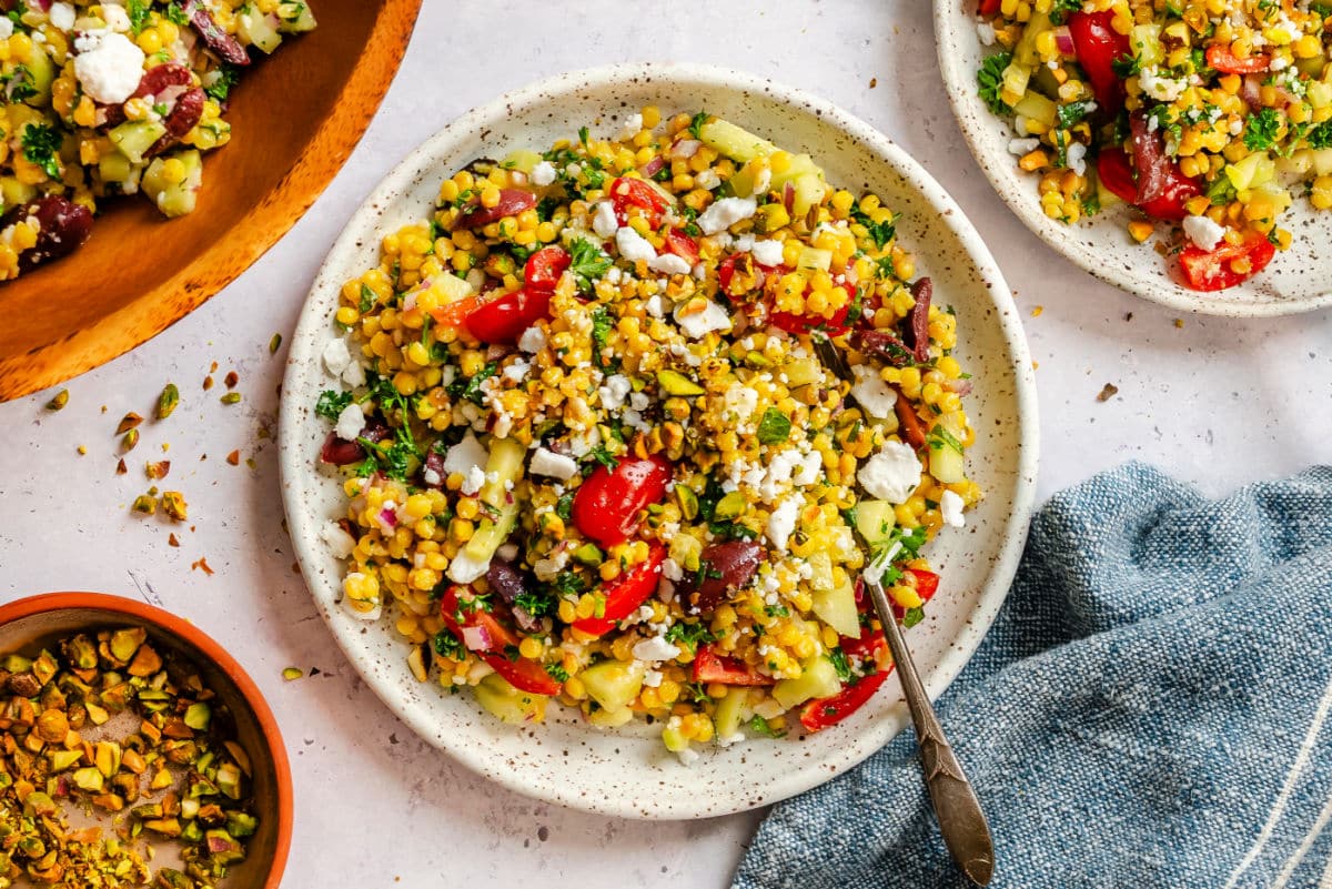A plate of pearl couscous salad next to a dish of chopped pistachios and a kitchen towel.