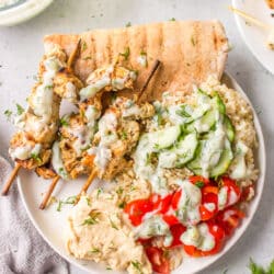 A plate with grilled Greek chicken skewers, cucumber, tomato, rice and pita topped with tzatziki.