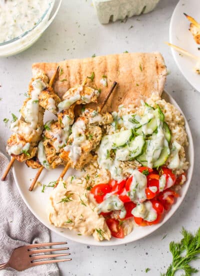 A plate with grilled Greek chicken skewers, cucumber, tomato, rice and pita topped with tzatziki.