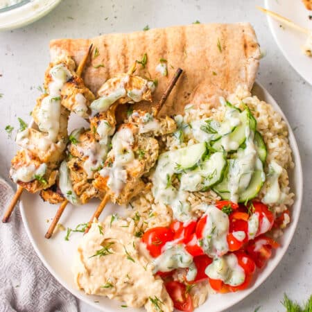 A plate with grilled Greek chicken skewers, cucumber, tomato, rice and pita topped with tzatziki.