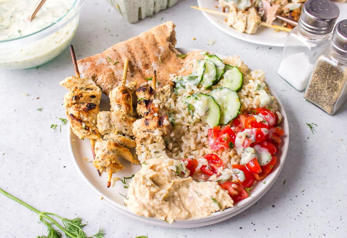 A plate of pita, Greek chicken skewers, sliced cucumber, and cut tomatoes next to a bowl of tzatziki.