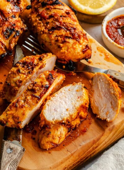 Cut harissa chicken on a wooden cutting board next to a dish of paste.