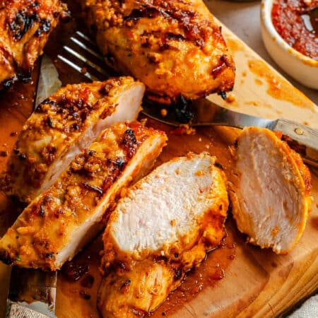 Cut harissa chicken on a wooden cutting board next to a dish of paste.