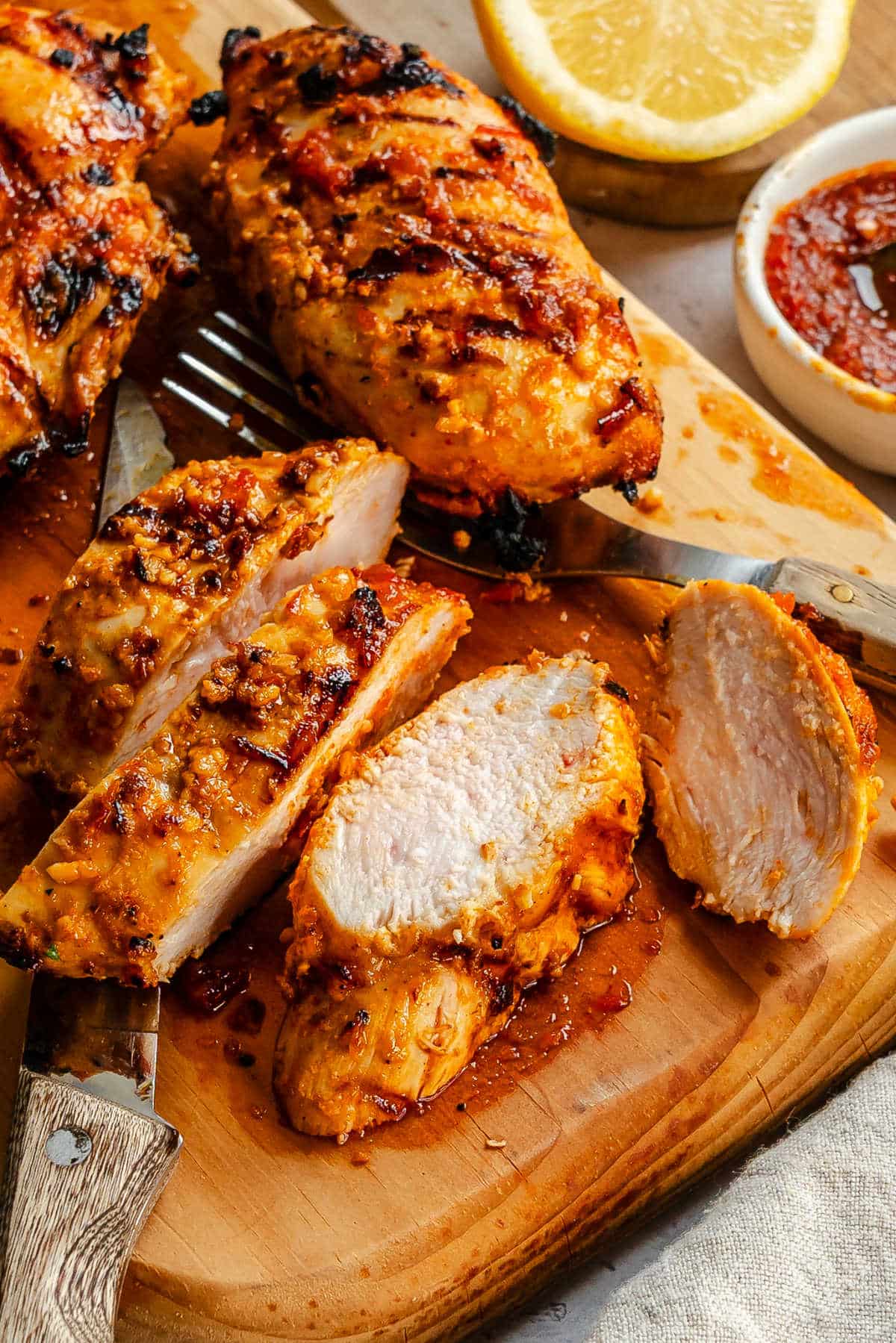 Cut harissa chicken on a wooden cutting board next to a dish of paste.