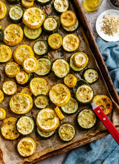 A sheet pan full of roasted summer squash and zucchini next to olive oil and a dish of seasoning.