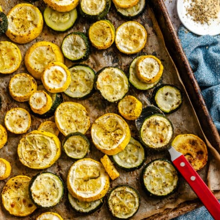 A sheet pan full of roasted summer squash and zucchini next to olive oil and a dish of seasoning.