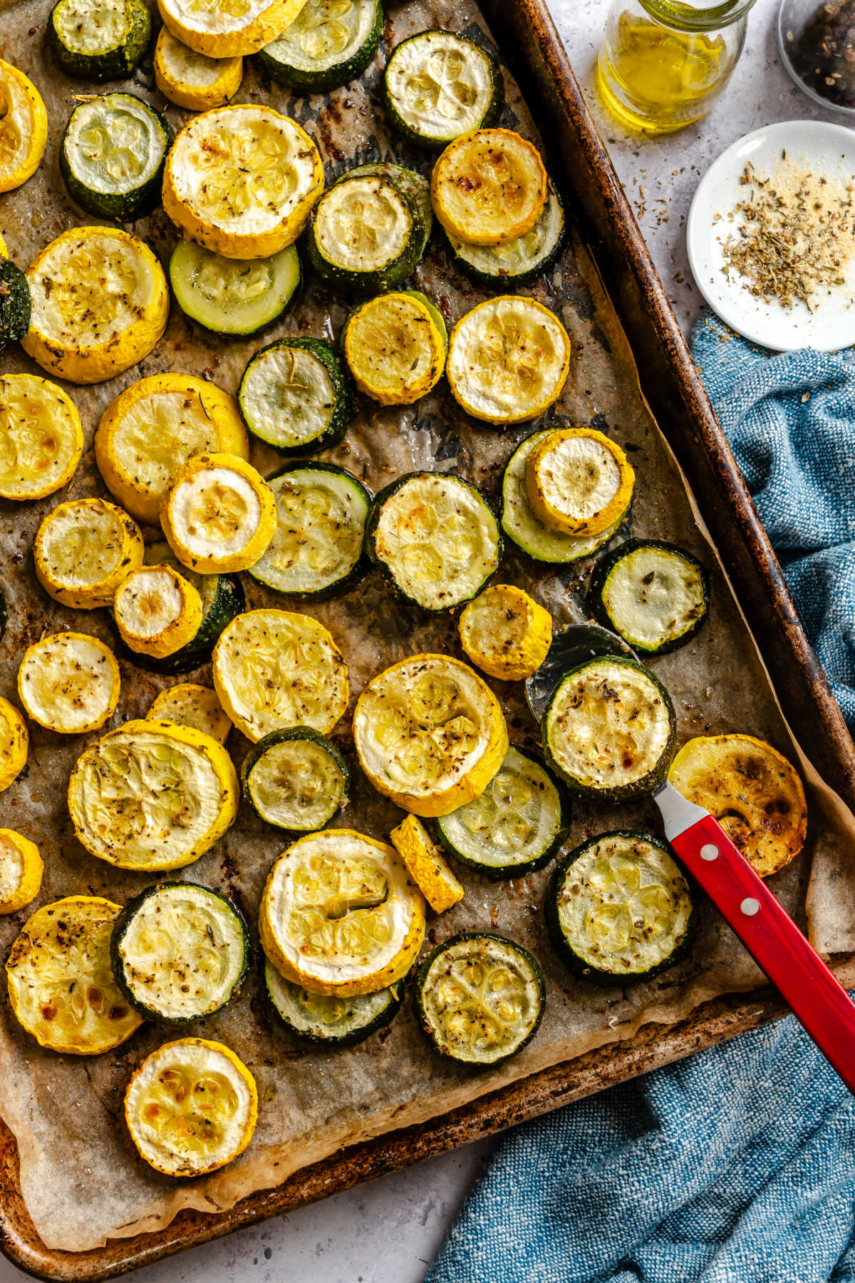 A sheet pan full of roasted summer squash and zucchini next to olive oil and a dish of seasoning.