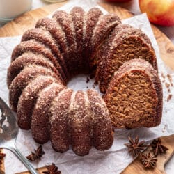 An apple cider cake with a slice sitting against it next to apples and a carafe of milk.