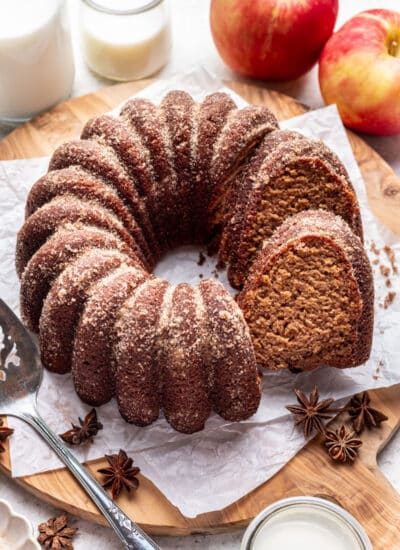 An apple cider cake with a slice sitting against it next to apples and a carafe of milk.