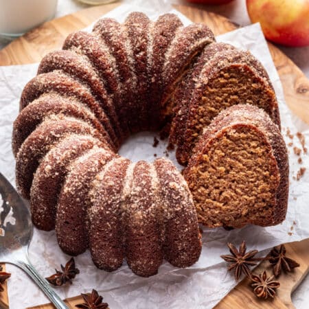 An apple cider cake with a slice sitting against it next to apples and a carafe of milk.