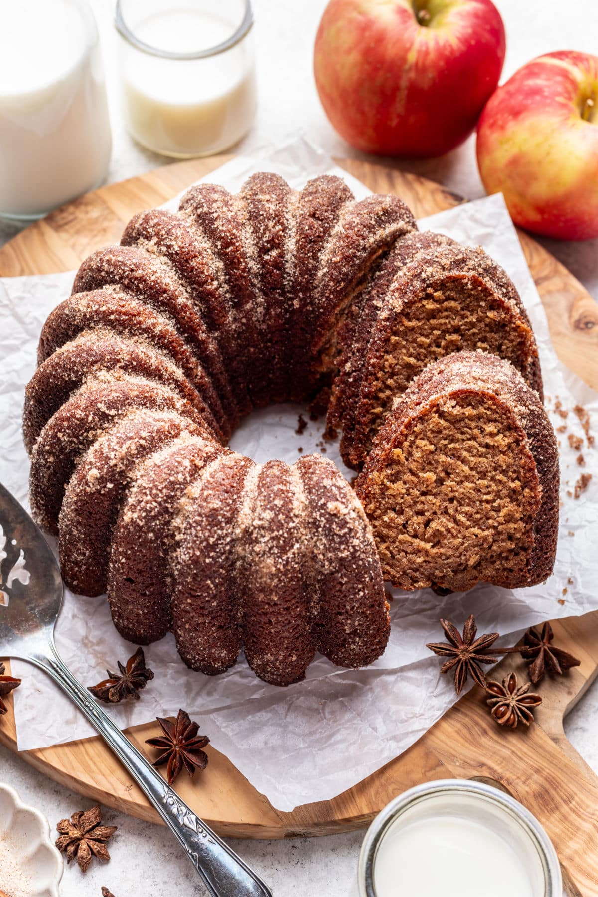 An apple cider cake with a slice sitting against it next to apples and a carafe of milk.