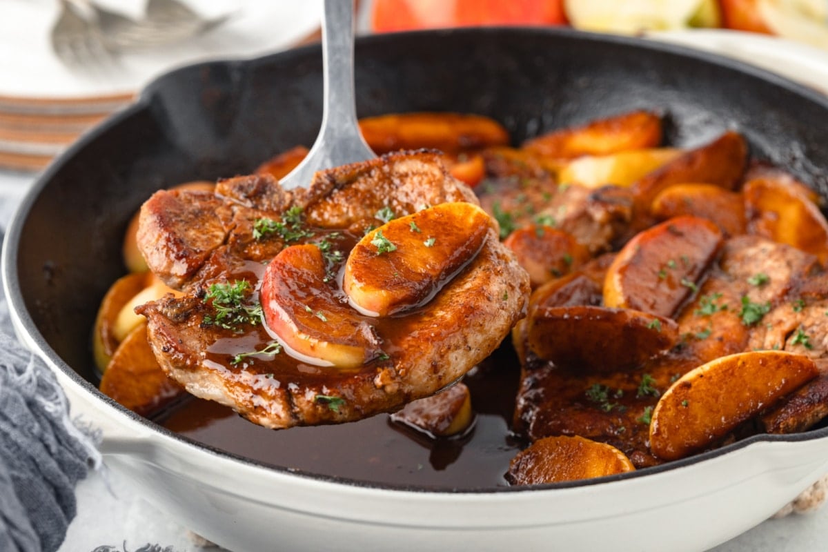A spatula lifting up a pork chop topped with sauteed apples.