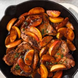 A skillet of pork chops and apples next to stacked plates.