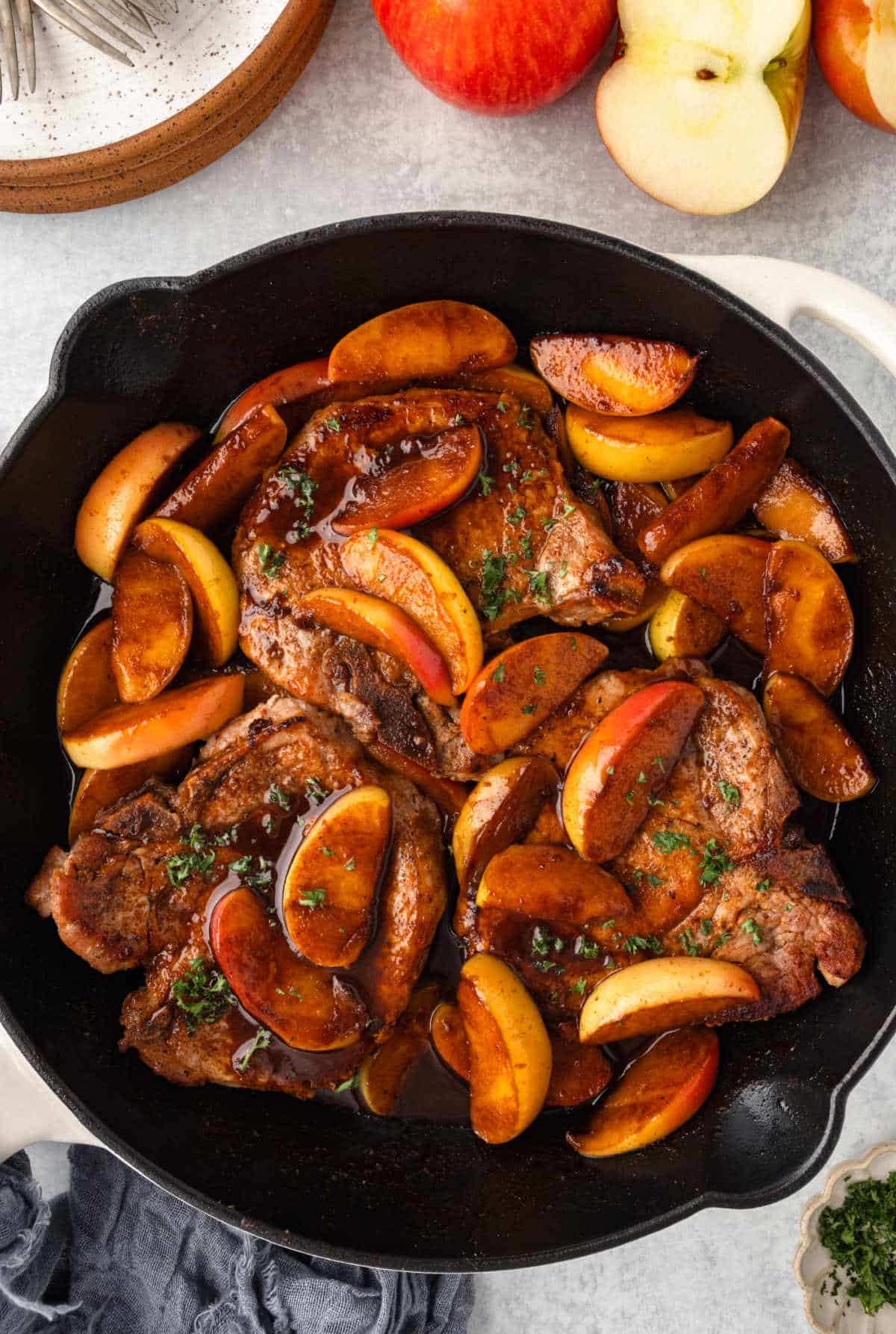 A skillet of pork chops and apples next to stacked plates.