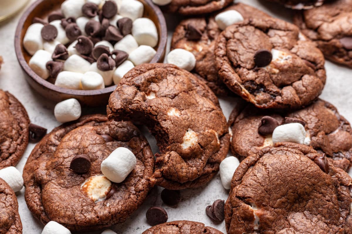 A chocolate marshmallow cookie with a bite missing at an angle against a dish of chocolate chips and marshmallows.
