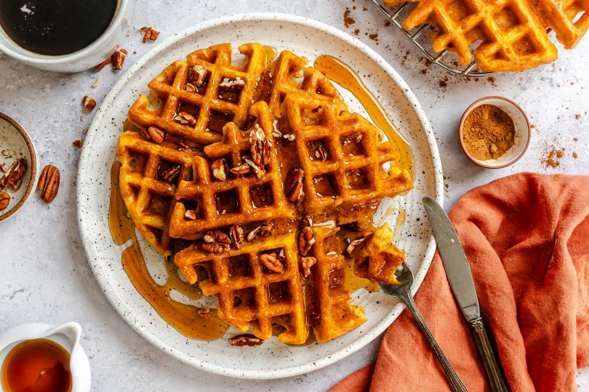 A fork holding a bite of pumpkin waffle next to two pumpkin waffles on a plate.